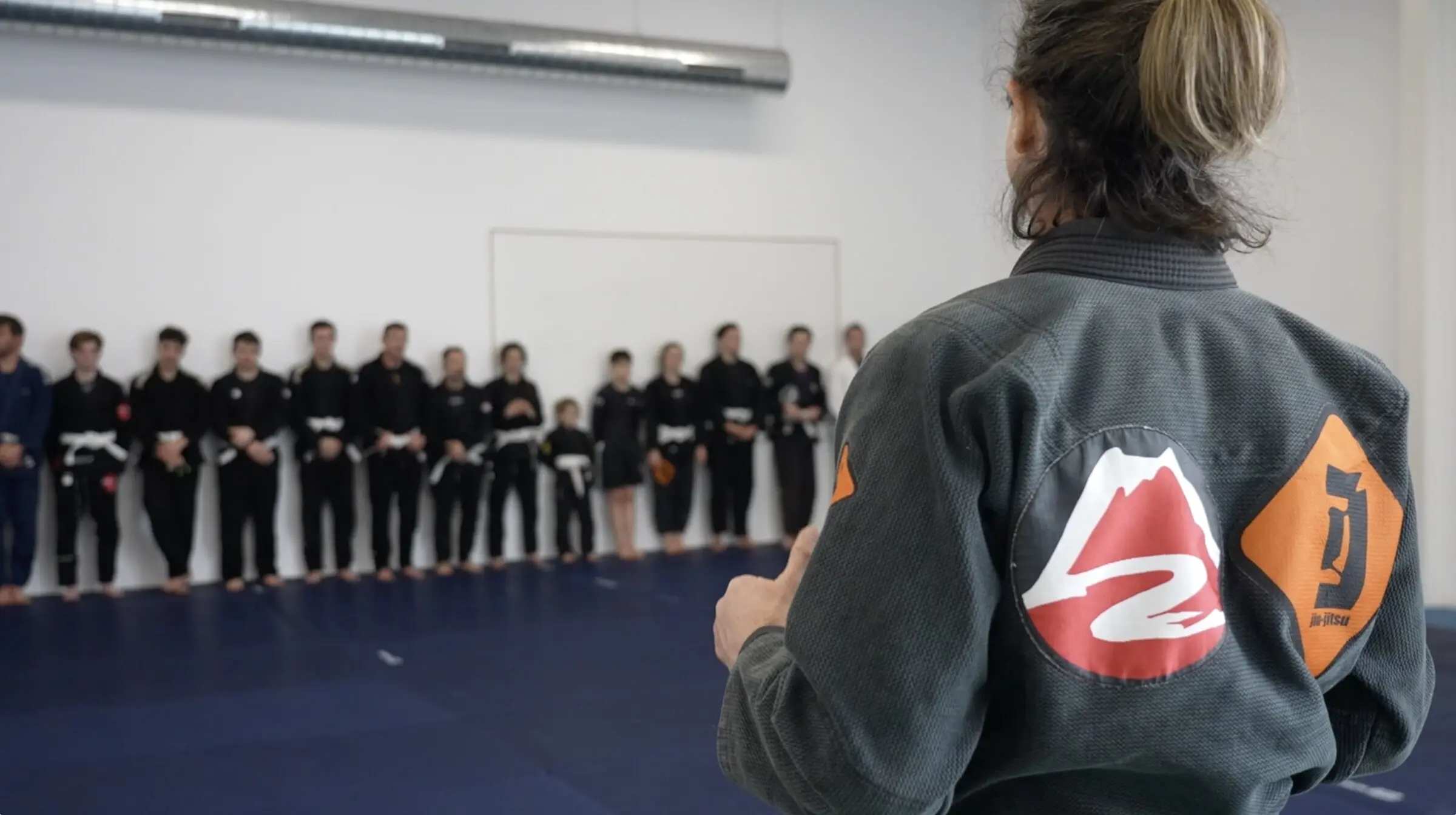 Sur le tatami, la professeure, dos à nous, l’écusson ICON sur l’épaule, fait face aux élèves alignés contre le mur